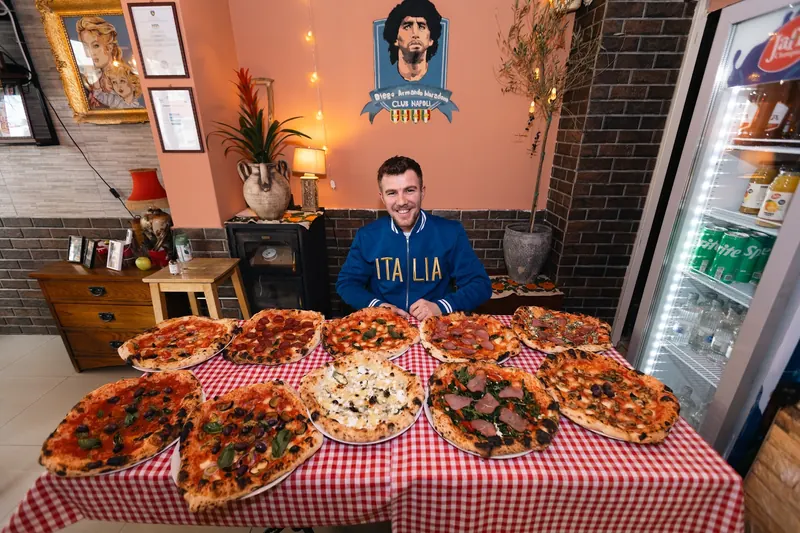 A pizzaiolo in an Italia jacket behind a red-checked table covered with ten wood-fired pizzas, under the Maradona Club Napoli mural